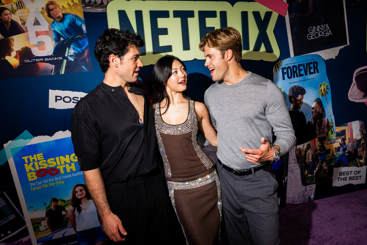 Three people posing together at a Netflix event, standing in front of a colorful backdrop with posters from various Netflix series and movies, engaging in conversation and smiling.