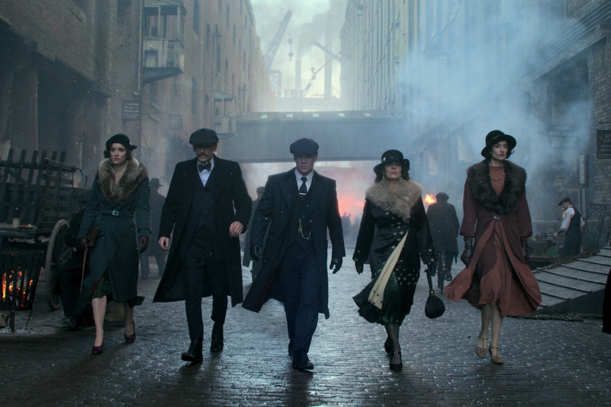 Five people in 1920s-style clothing walk confidently down a cobblestone street in an industrial city setting, surrounded by brick buildings, smoke, and period details.