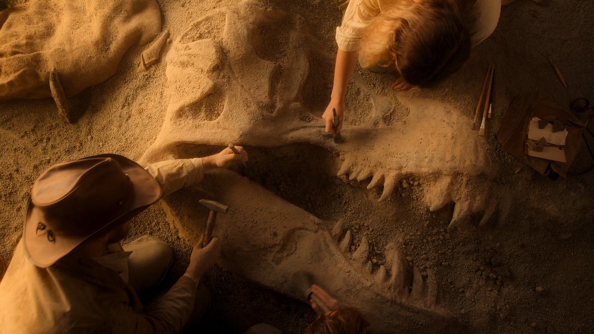 Two people carefully excavating a large dinosaur skull fossil in sandy soil, using small brushes and tools, with excavation equipment nearby, in a warm, softly lit dig site environment.