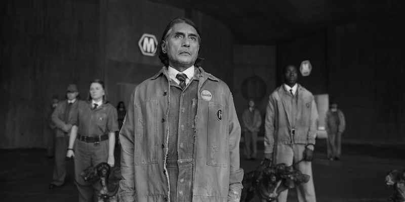 Black and white image of Ramon Tikaram as Charon wearing a worn out jumpsuit and jacket in 'KAOS'