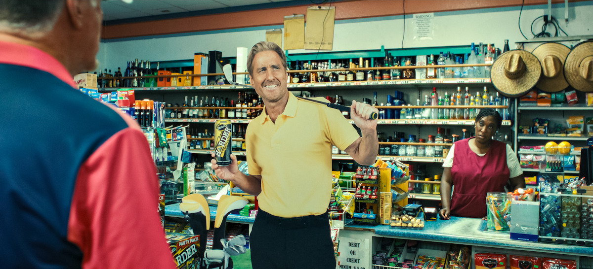 Man in yellow shirt holding a drink and gesturing in a colorful convenience store, another man in red shirt foreground, and store clerk behind counter. Shelves are stocked with snacks, drinks, and sombreros on wall.