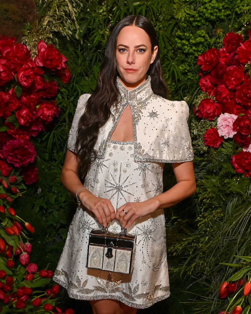 Woman in a white embellished dress with silver accents and long dark hair, standing in front of lush green foliage and vibrant red flowers, holding a small decorative handbag, posing at a formal event.