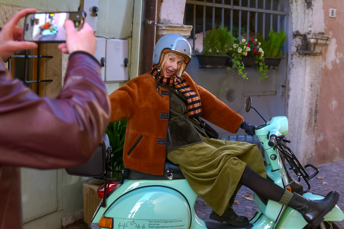 Woman in helmet and scarf poses playfully on a vintage light blue scooter while someone takes her photo in a charming European alley with plants and cobblestone.