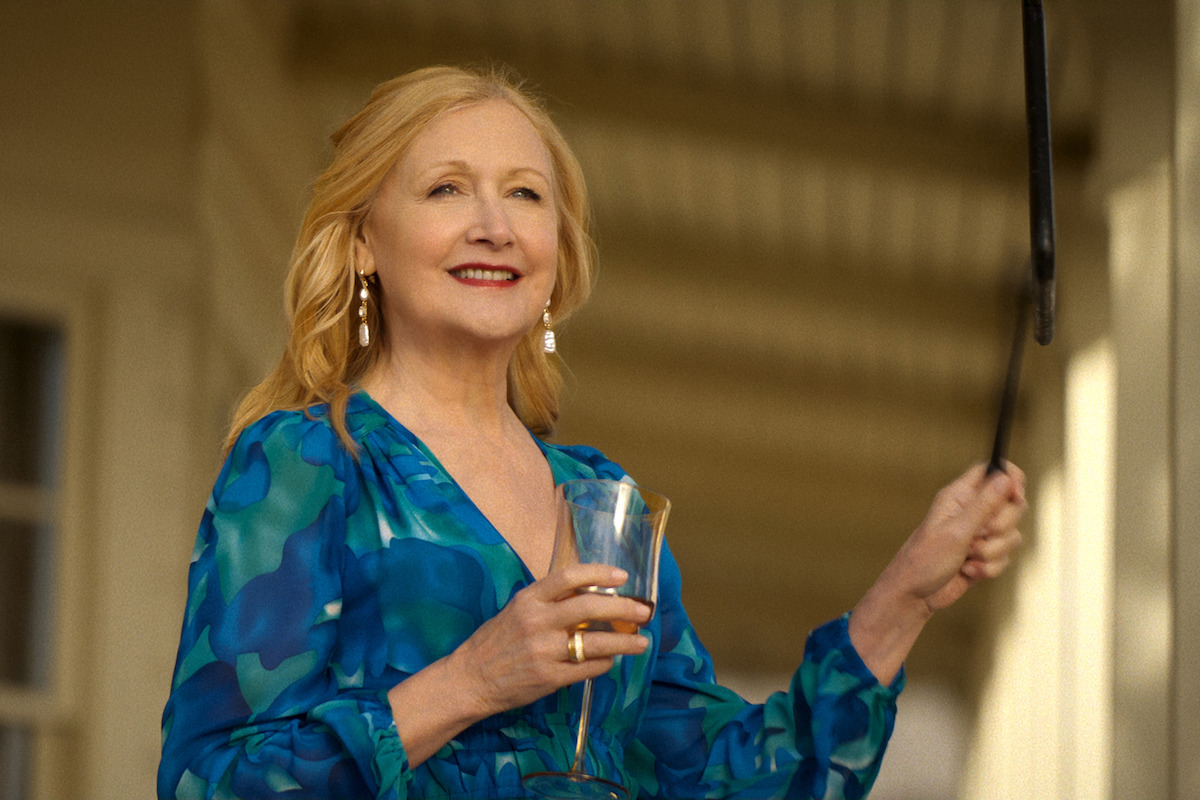 Woman in a blue patterned dress stands on a porch holding a drink, smiling, with sunlight illuminating her and a window and railing visible in the background.