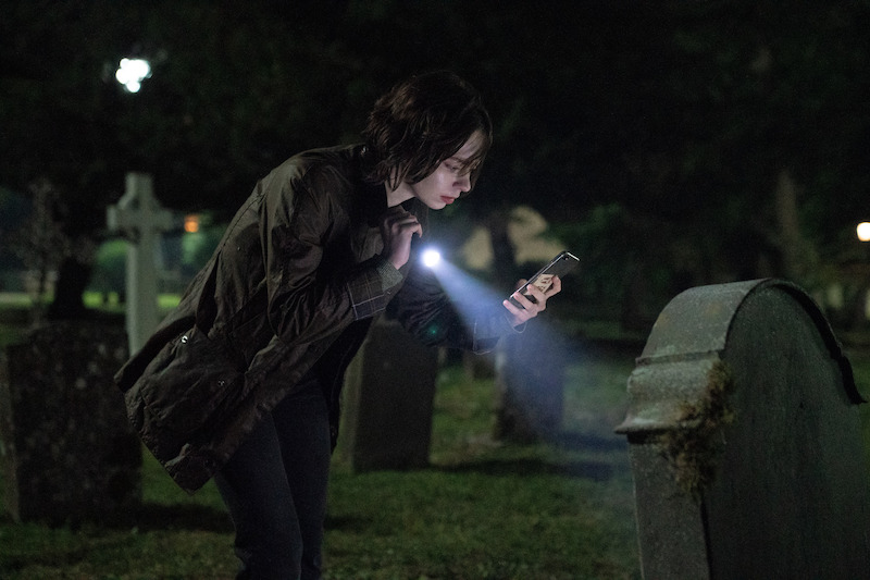 Person in dark clothing examines a gravestone at night in a cemetery, using the flashlight on their phone to illuminate the scene; the background is dimly lit with other gravestones and trees.