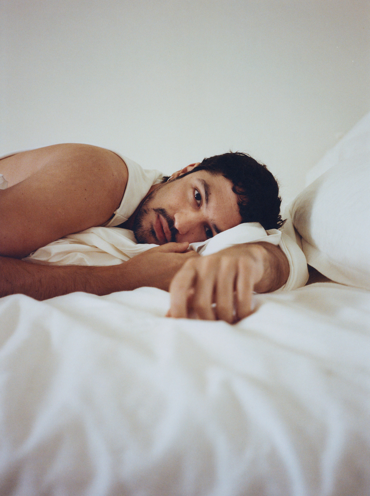 Gabriel Leone lies in a white bed with a sultry expression.