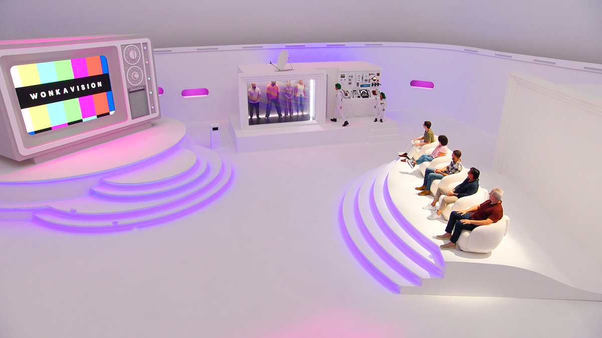 Futuristic white studio with a large retro TV screen, contestants seated in modern chairs, and participants standing in a glass enclosure; ambient pink and purple lighting.