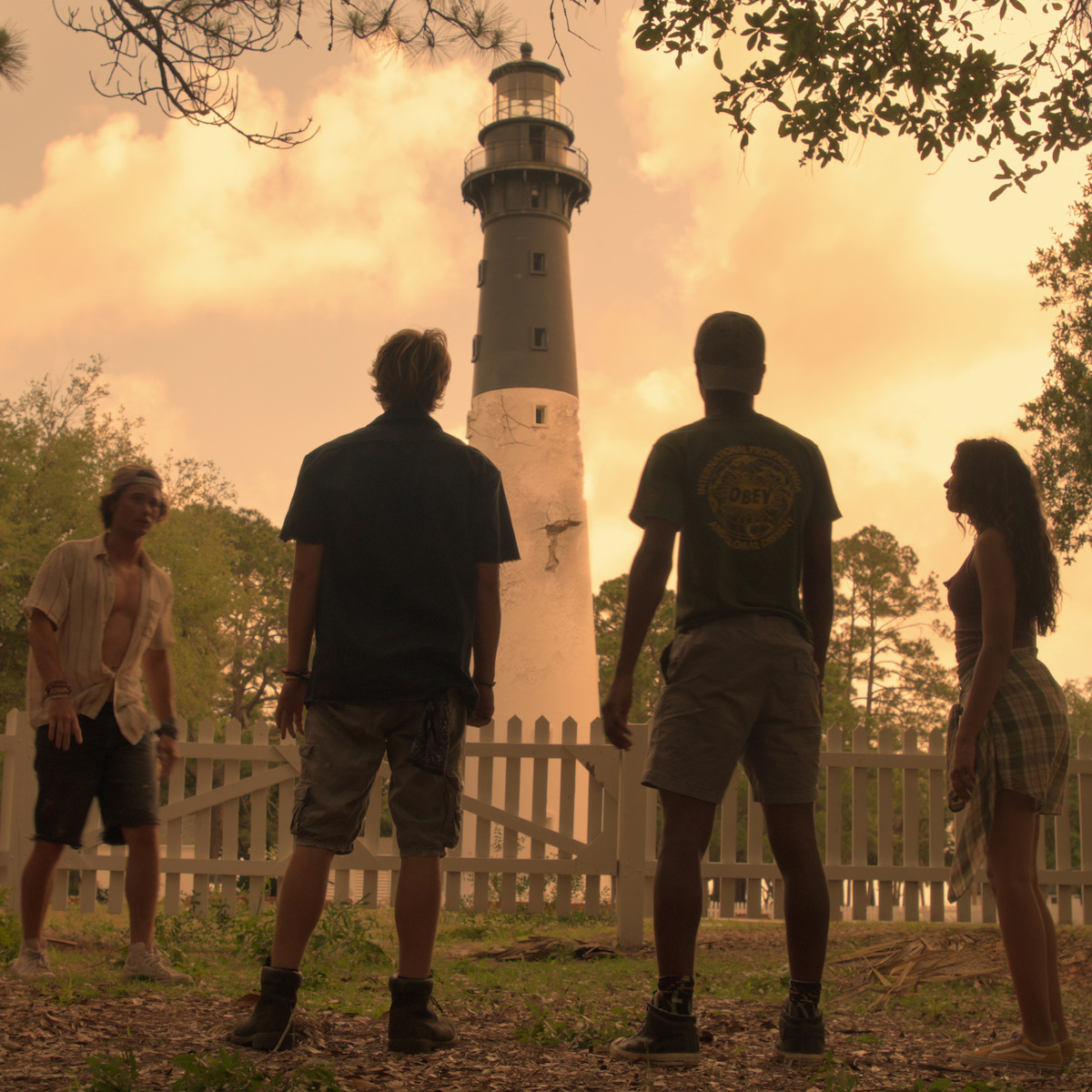 Quatro pessoas estão em pé em frente a um farol alto cercado por uma cerca branca, com árvores e um céu nublado e aconchegante ao fundo, dando à cena um clima de verão e aventura.