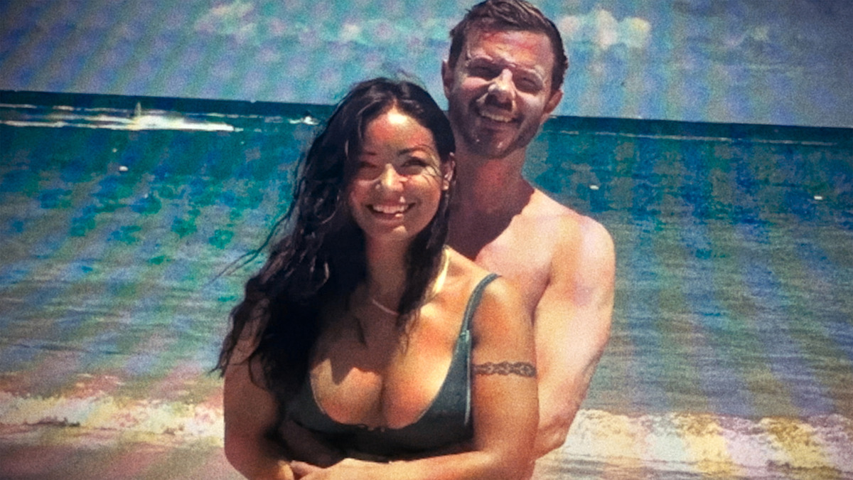 A smiling couple standing together on a sunny beach with waves and a blue ocean in the background.