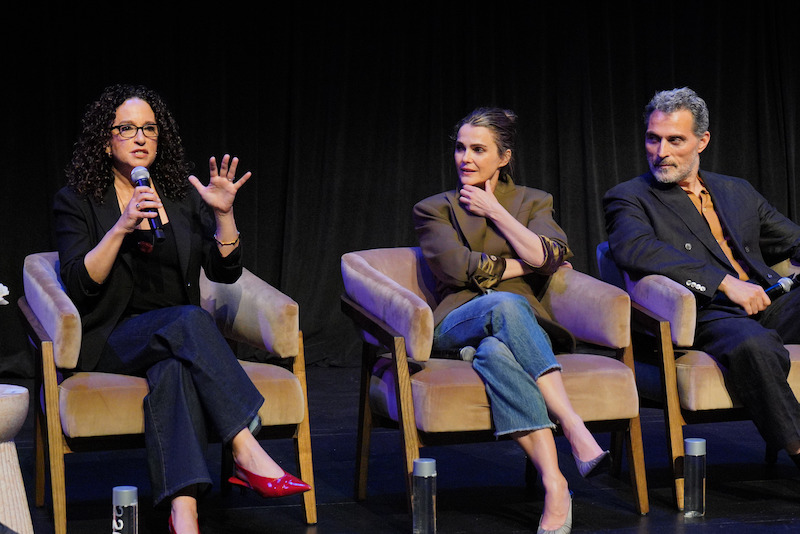 Three people sit on tan chairs on a stage with black curtains. The woman on the left speaks into a microphone, gesturing with her hand, while the other two panelists listen attentively. Bottled water is placed in front of each chair.