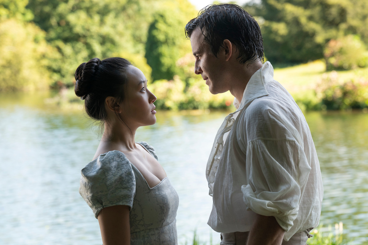 Two people in period clothing stand facing each other by a lake, surrounded by greenery and trees, in an outdoor romantic setting.
