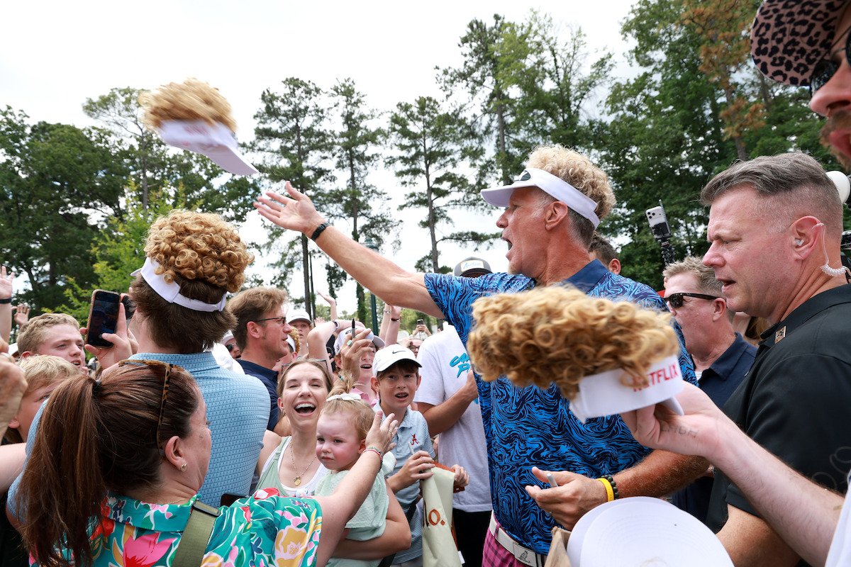 A man in a blue shirt throws novelty visors with curly wigs to a crowd of smiling people, including families and children, at an outdoor event surrounded by trees on a sunny day.