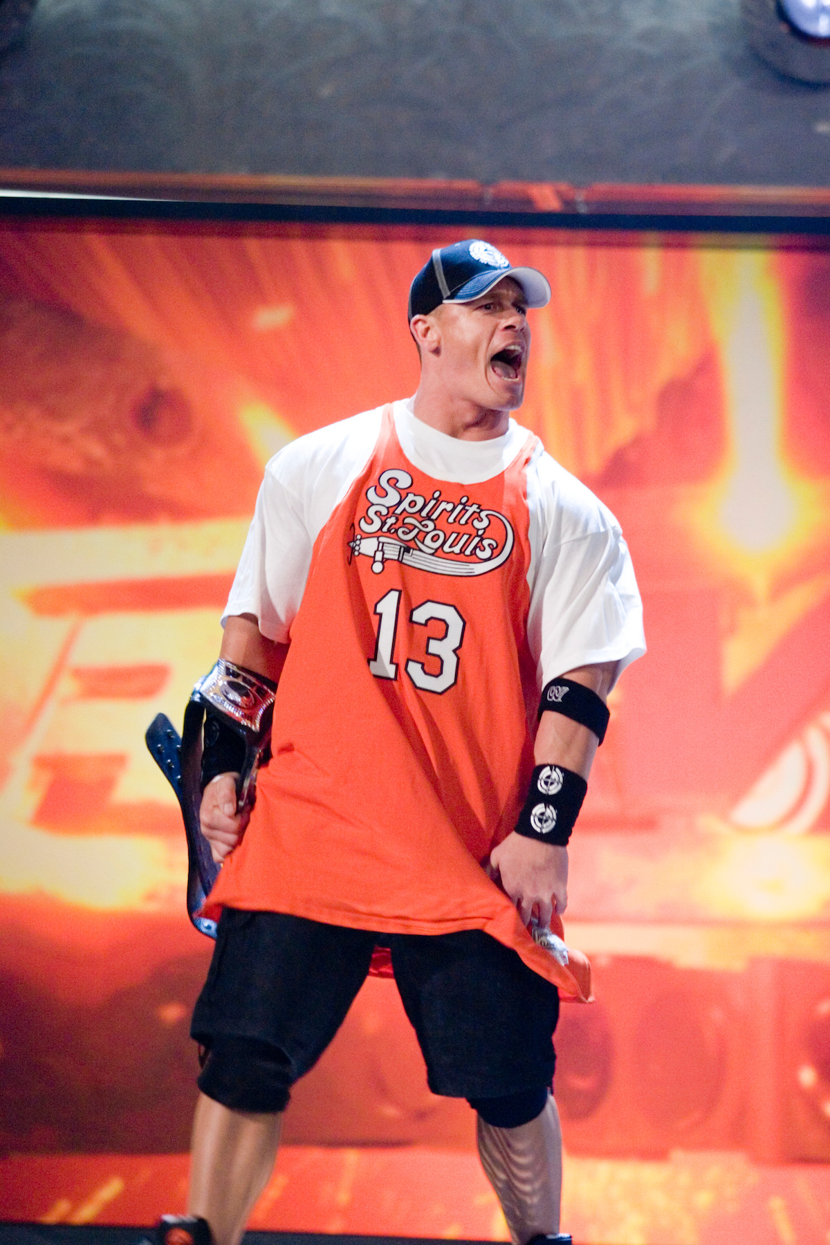 A muscular man in sports attire and a cap passionately poses on a vibrant stage with energetic lighting, conveying excitement and intensity, likely in a performance or competition setting.