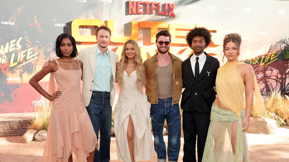 Six people pose together on a red carpet at an outdoor Netflix premiere event, dressed in fashionable attire, standing in front of a themed backdrop with desert elements and promotional text.