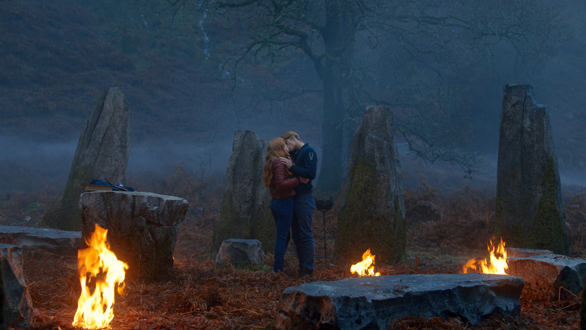 A couple embraces in a misty stone circle at night, surrounded by flames and large standing stones, with a dark, foggy forest in the background.