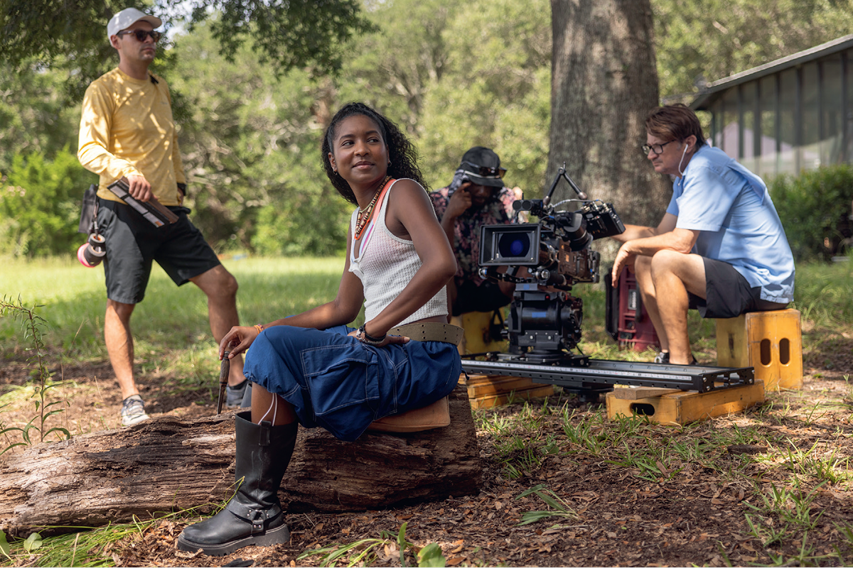 Woman sits on a log outdoors with a film crew, including a camera operator and crew members with equipment, surrounded by trees and grass, during a film shoot in a natural, sunny environment.