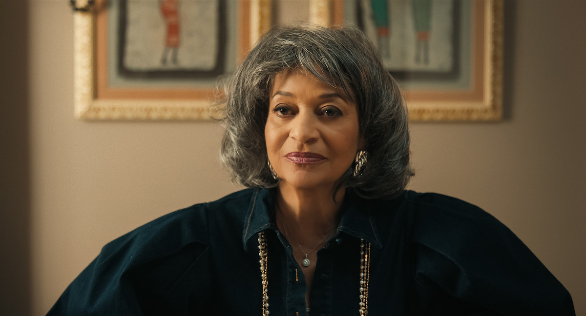 Woman with gray hair wearing a dark blouse and gold necklace, sitting indoors in front of framed artwork on a beige wall, softly lit room.