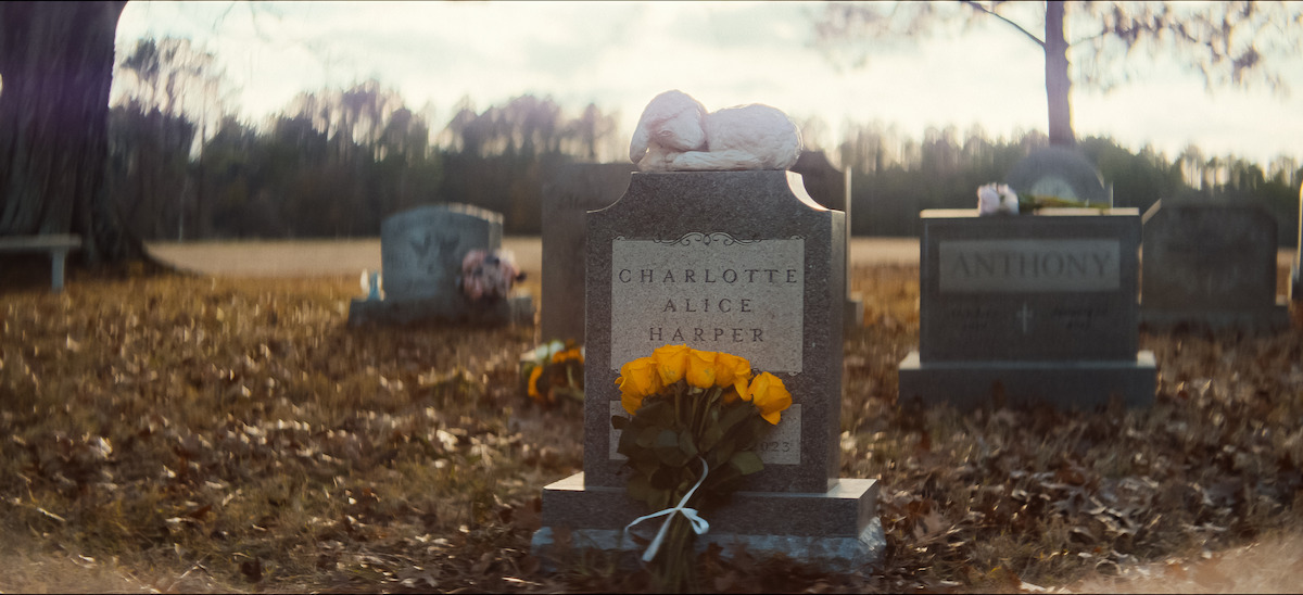 Gravestone with engraved text that reads "Charlotte Alice Harper" with yellow flowers and a stuffed animal on top in a quiet, leaf-covered cemetery surrounded by bare trees.