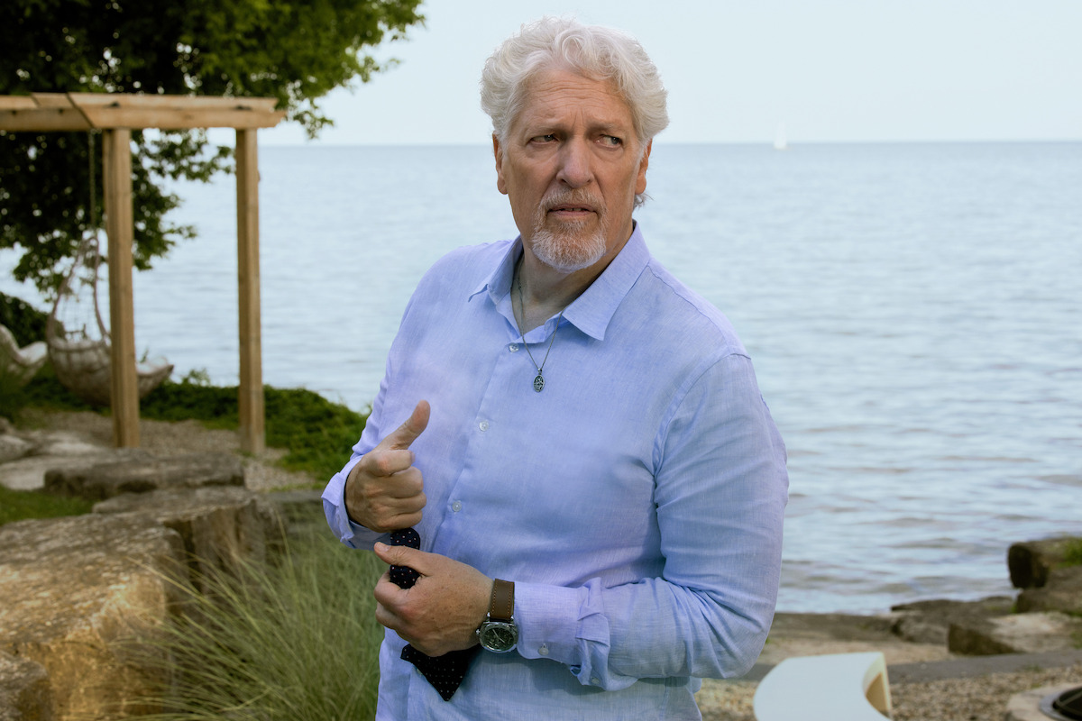 Older man with white hair and beard stands outdoors by a lakeshore, wearing a light blue shirt and holding a necktie. Natural background with rocks, trees, and water on a clear day.
