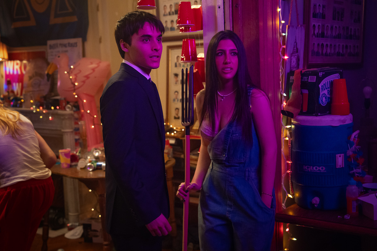 A young man in a suit and a young woman in overalls stand together at a lively, colorful house party decorated with string lights, red cups, and party supplies, looking surprised or concerned.