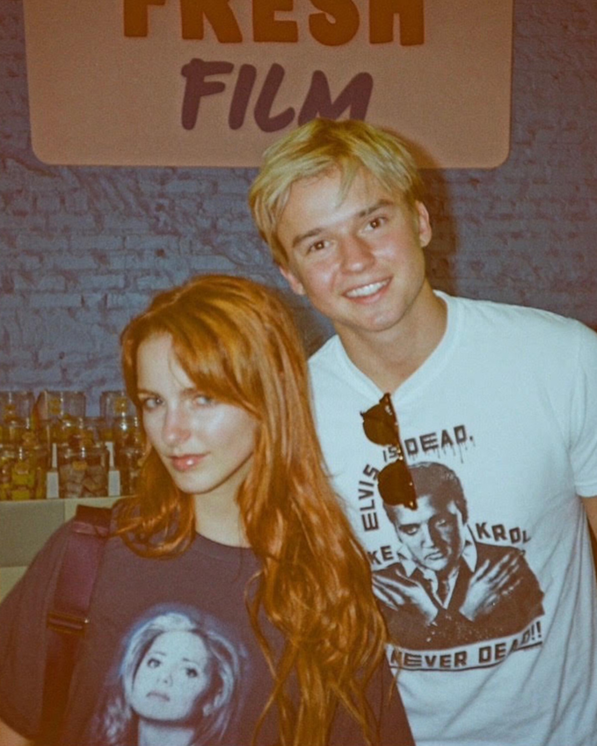 Two young adults in graphic t-shirts stand together and smile inside a shop with jars on shelves and a sign reading "FRESH FILM" on a blue brick wall in the background.
