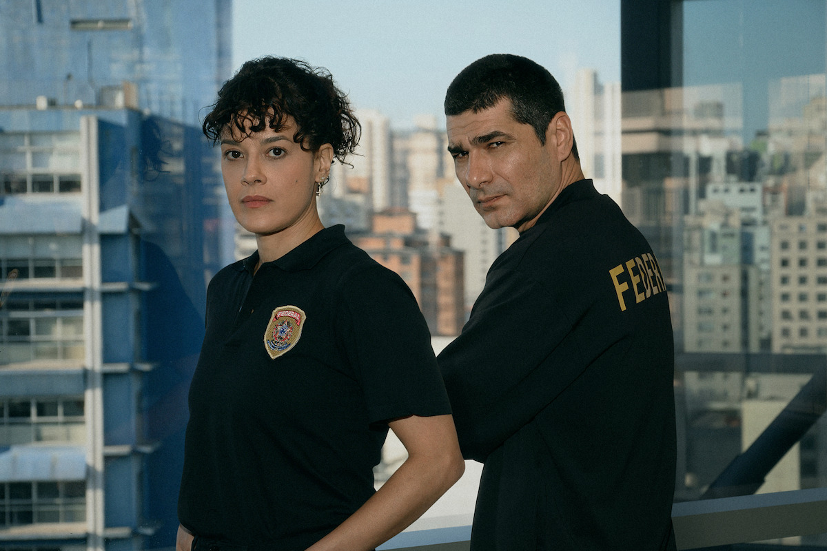 Two individuals in black uniforms, one with a police badge and the other with "Federal" on their back, standing in front of a window overlooking a city skyline.