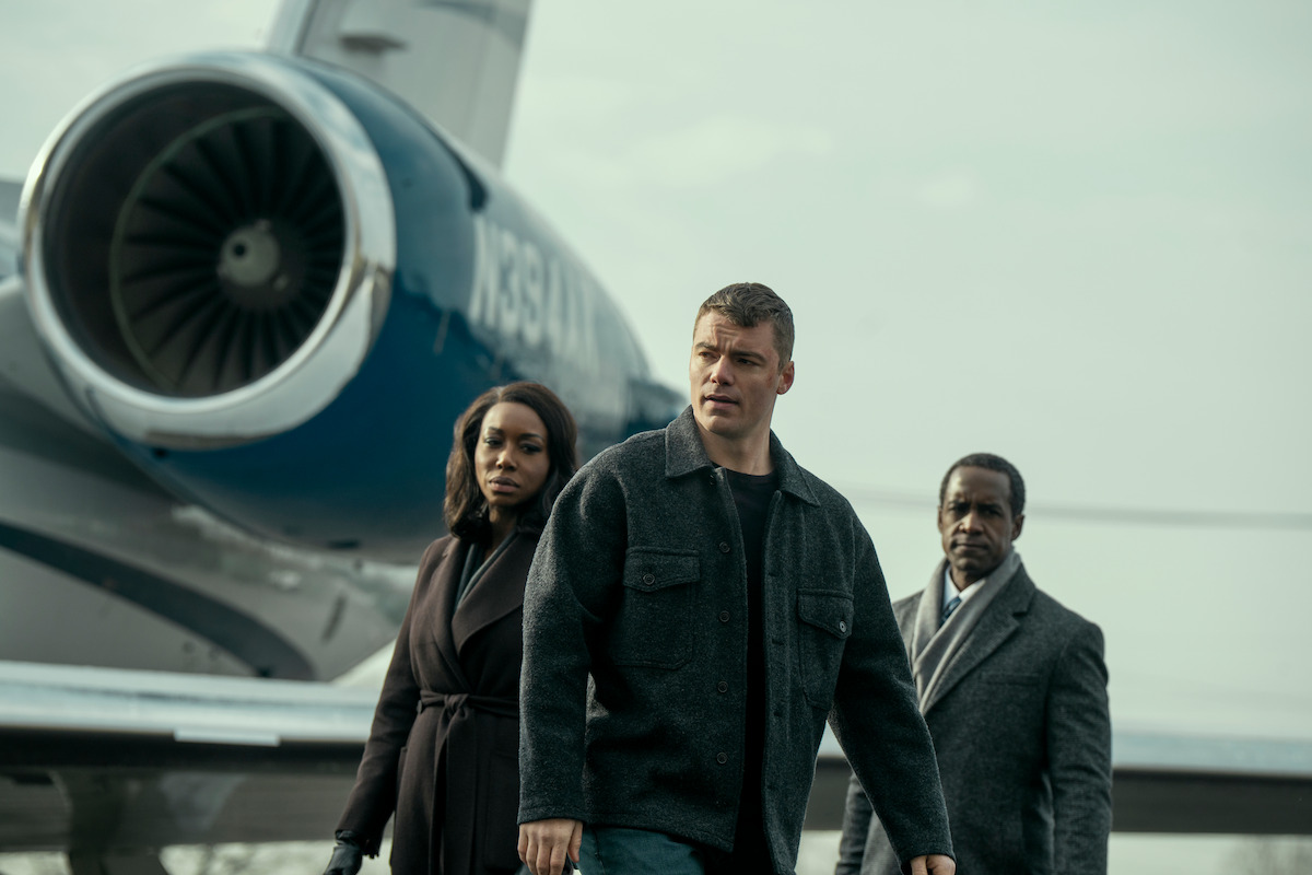 Three people stand near a jet on an airport runway, looking serious and focused. The group is dressed in dark clothing, and the environment appears tense and overcast.