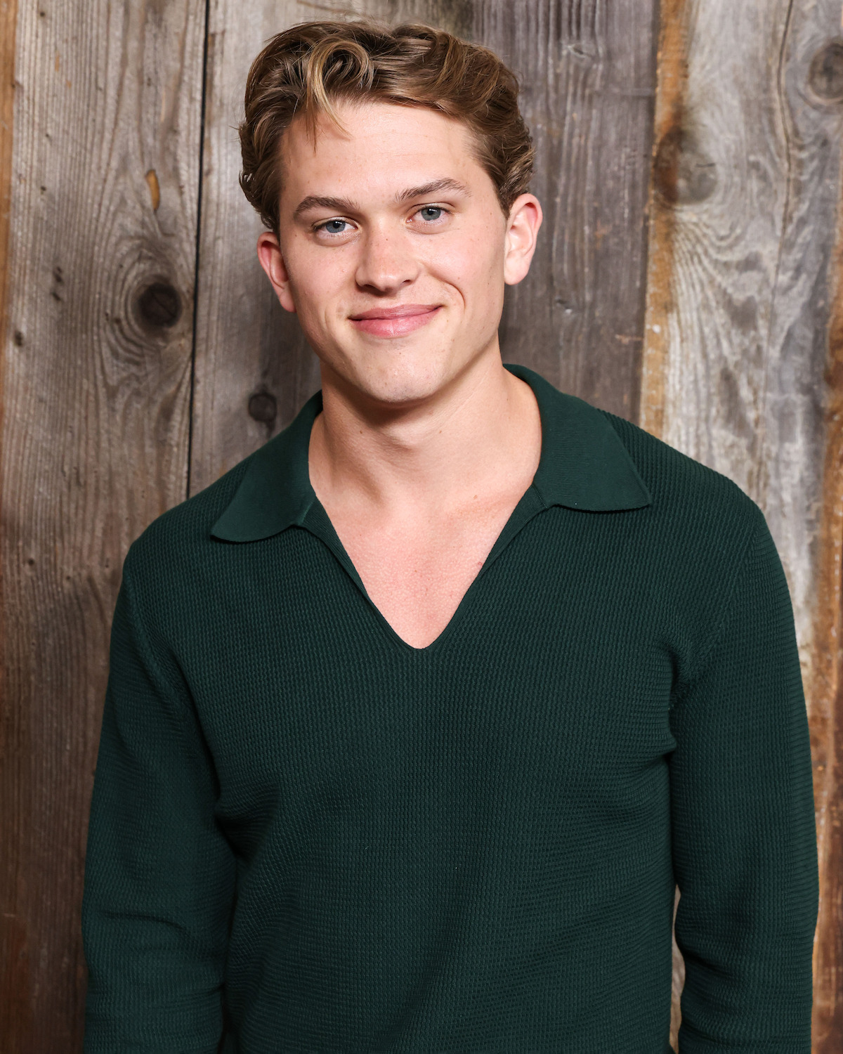Young man with short light brown hair wearing a dark green collared shirt, standing in front of a rustic wooden wall, smiling at the camera in a casual indoor setting.