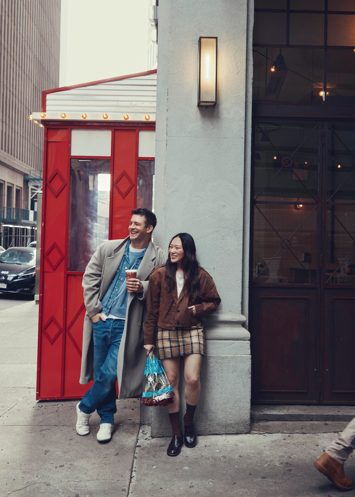 A man and woman stand on a city sidewalk, leaning against a building corner, smiling and holding drinks. They are dressed in casual, stylish clothing. There is a red booth and modern city buildings in the background.