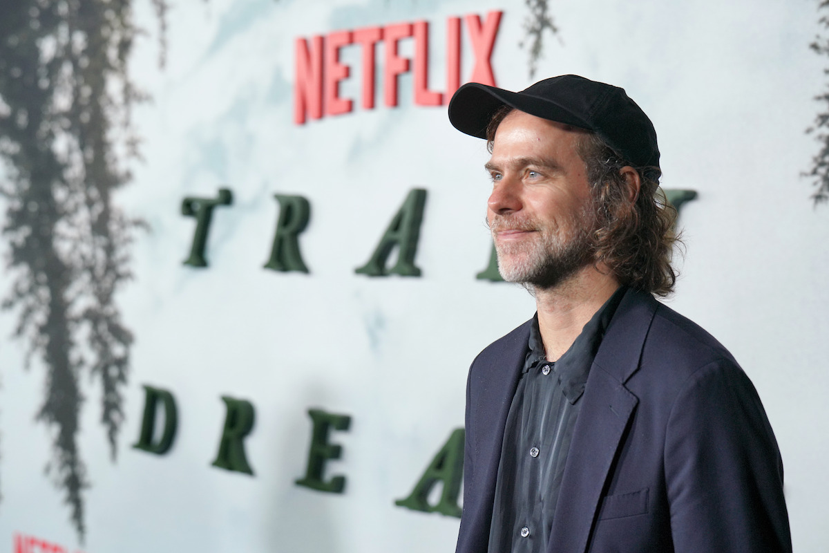 A man in a black cap and suit stands smiling at a Netflix event with a nature-inspired backdrop, creating a casual and positive atmosphere.
