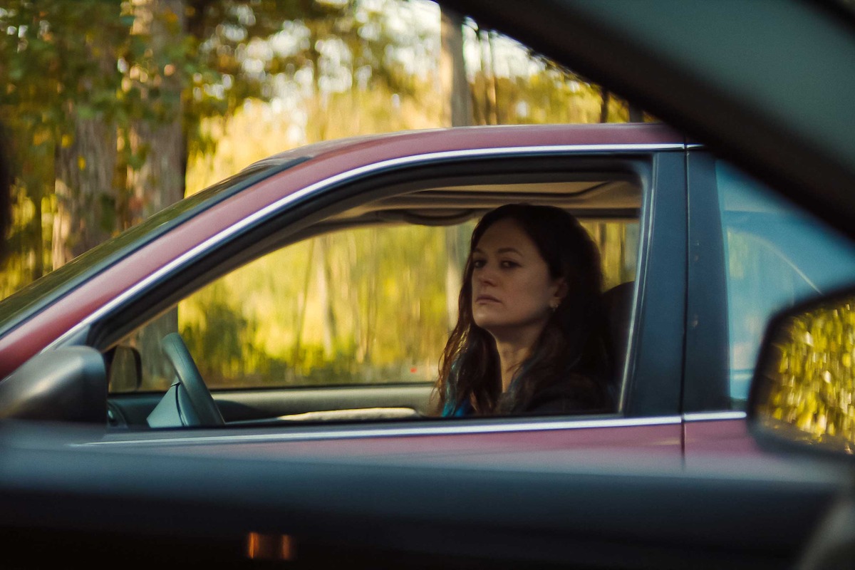 Woman with long dark hair sits in the driver's seat of a red car, looking slightly serious. The photo is taken from another vehicle, with trees and greenery visible outside the window, suggesting a daytime outdoor setting.