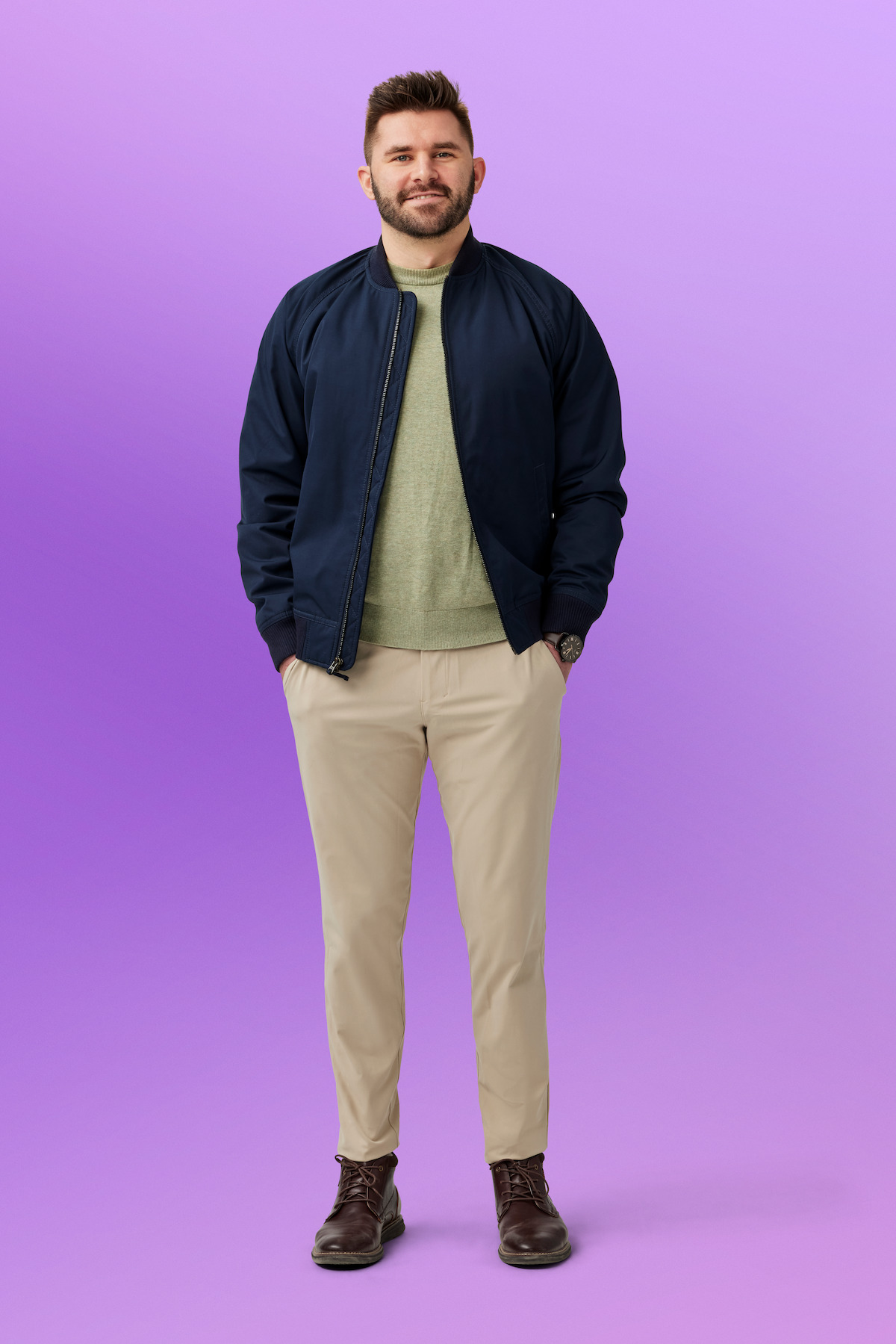 Connor standing with hands in pockets, wearing a dark jacket, light sweater, beige pants, and brown shoes, against a purple studio background.