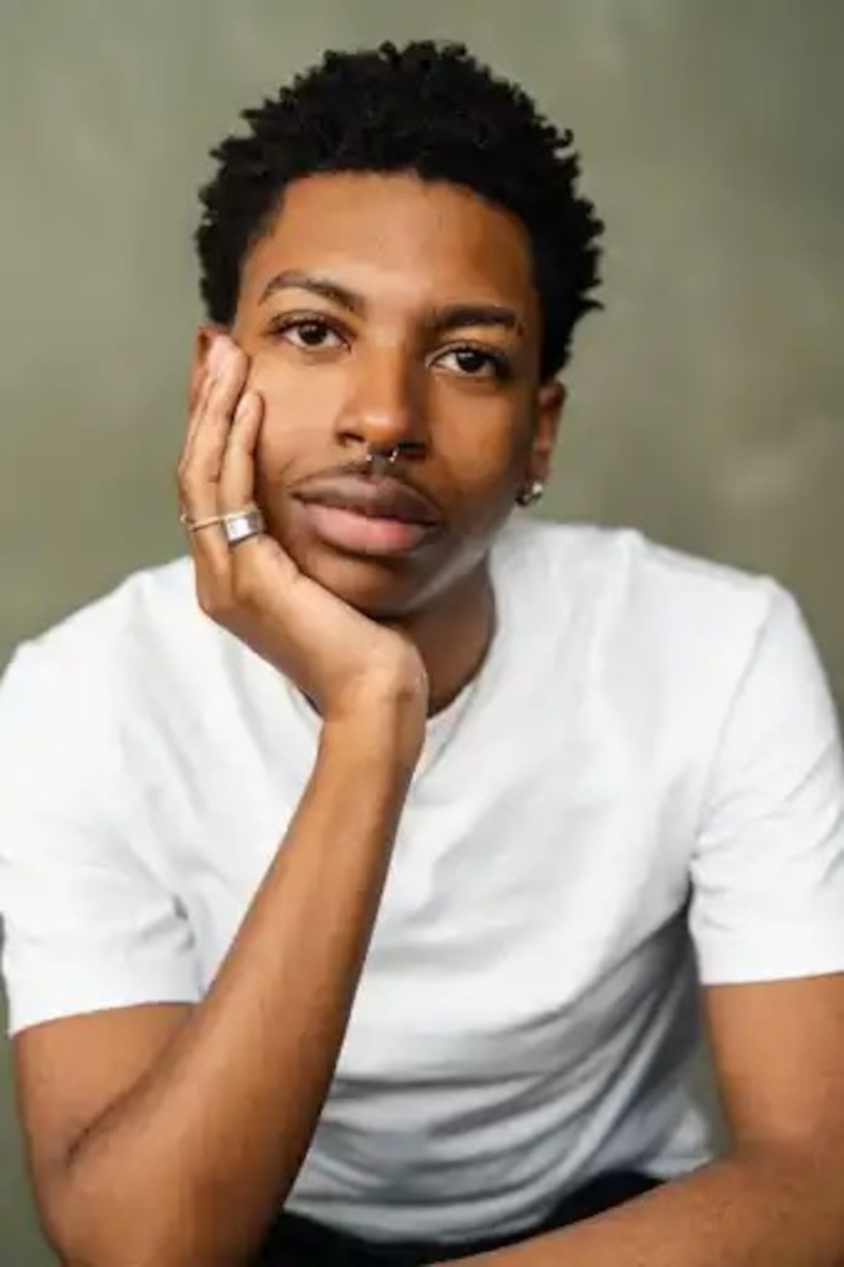 Man wearing a sleeveless white sweater, standing against a plain, neutral background, looking confidently at the camera under soft studio lighting.