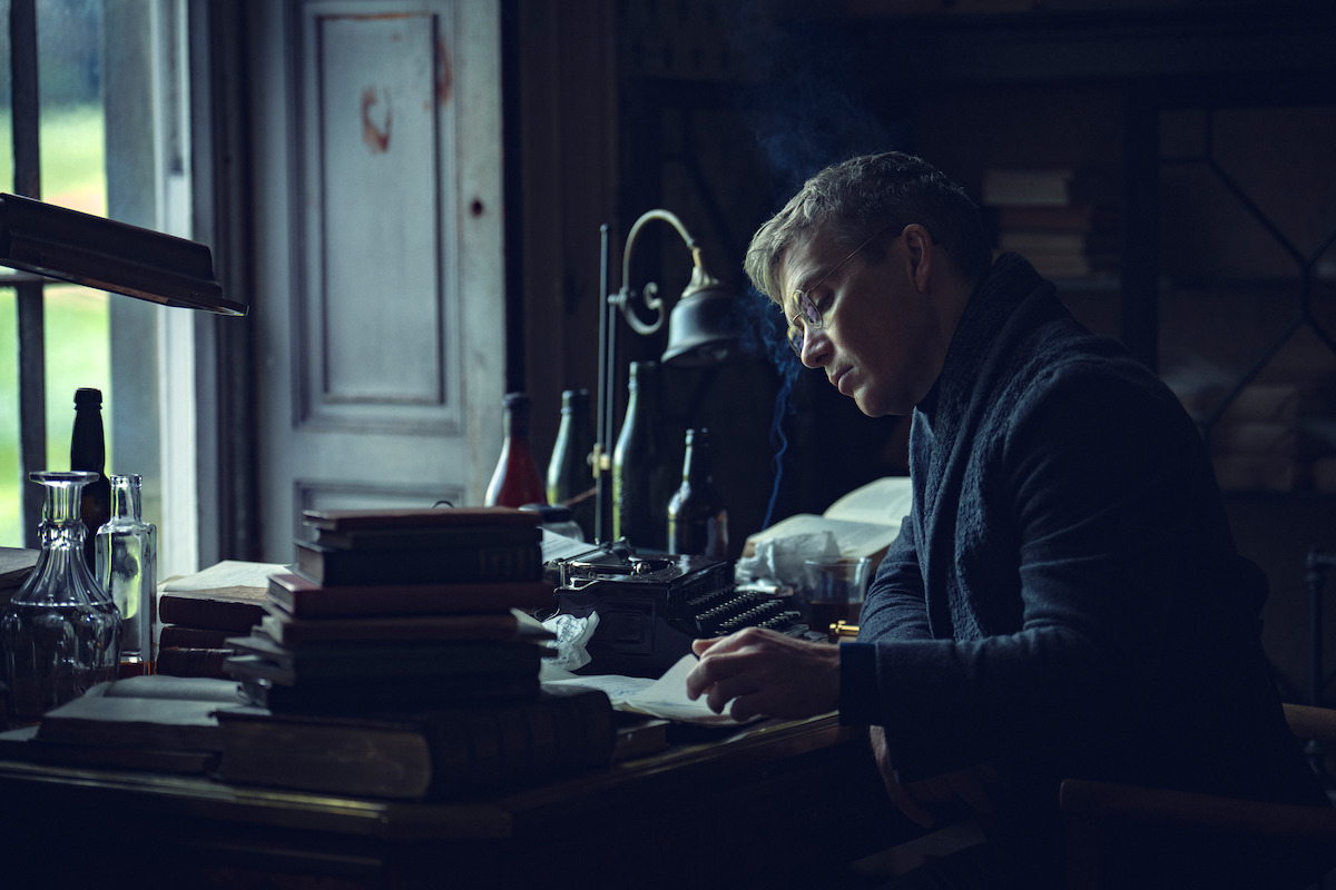 Man writing at a cluttered desk by a window in a dimly lit study, surrounded by stacks of books, bottles, a typewriter, and old-fashioned decor, creating a contemplative and vintage atmosphere.