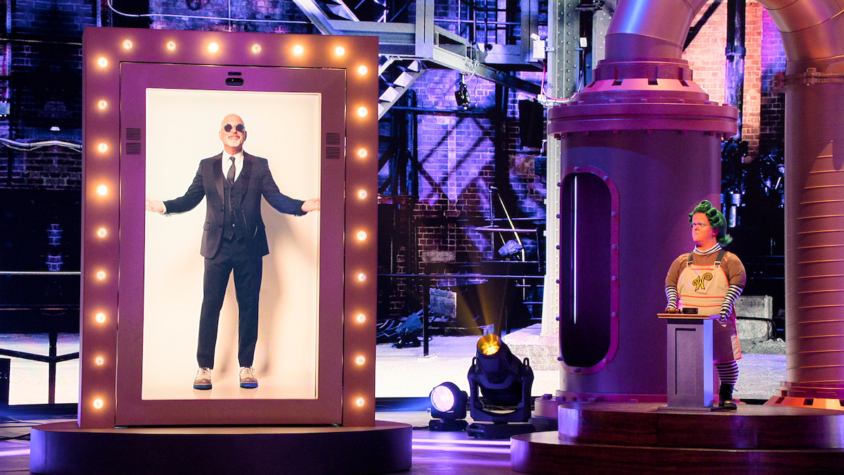 Bald man in a suit stands in a large, illuminated frame on a stage; a person in colorful costume operates machinery in a whimsical, industrial-themed set with purple lighting.