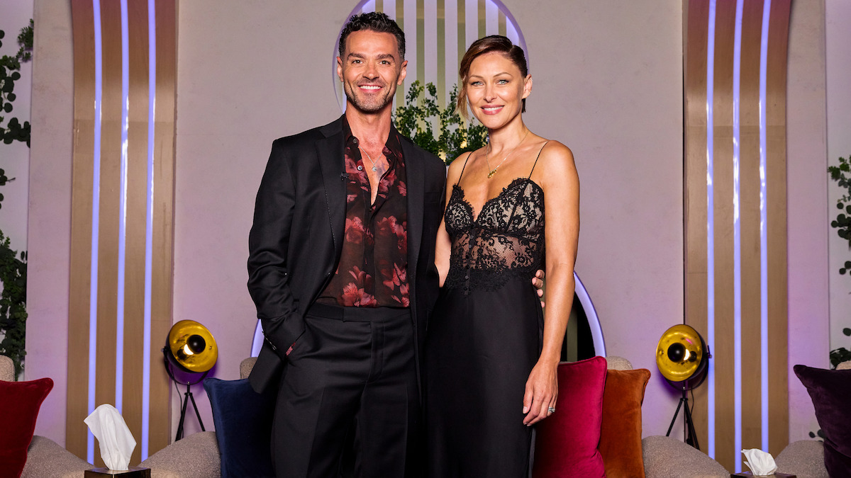 A man in a black suit and a woman in a black lace dress stand together smiling in a modern lounge with colorful cushions, round lights, plants, and decorative vertical lines in the background.