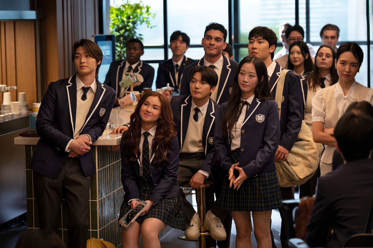 A group of high school students in uniforms gather indoors, some standing, some sitting, in a modern, well-lit room with large windows and plants, appearing attentive and relaxed, likely listening to someone speak.