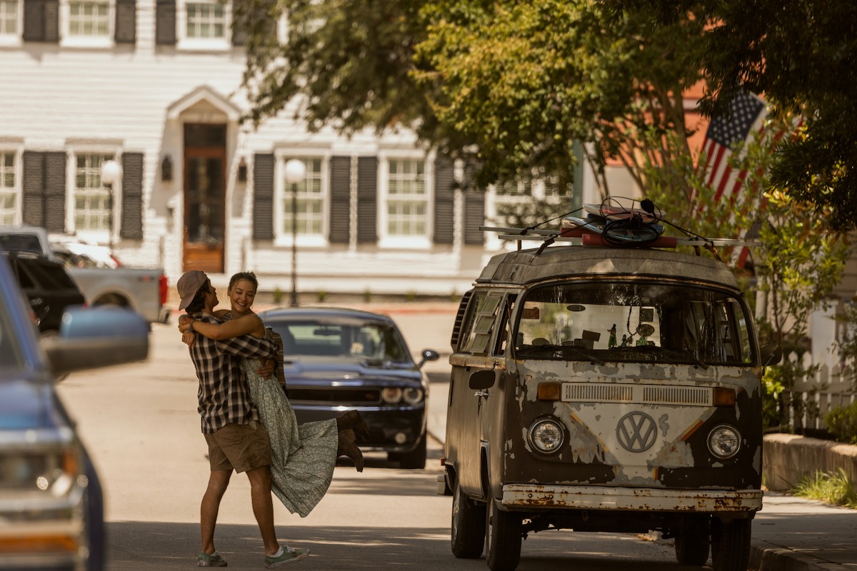 A man lifts a woman joyfully in a sunny residential street with parked cars and an old Volkswagen van with surfboards, American flag in background, trees and historic houses nearby.