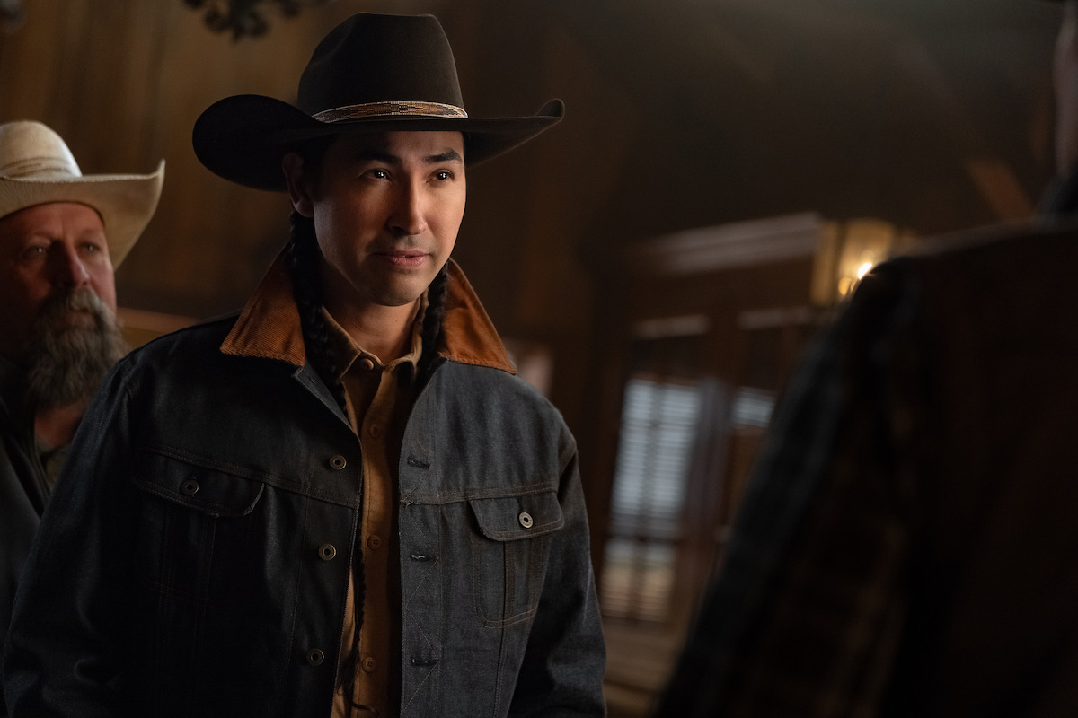 A man wearing a cowboy hat and denim jacket stands indoors in a warmly lit rustic room with wood paneling, accompanied by another man in a hat. The setting suggests a western or ranch environment.
