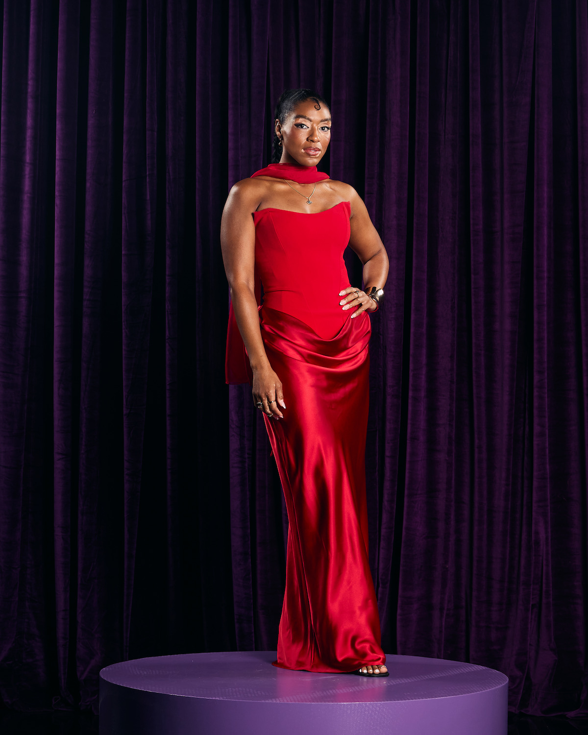 Woman in a red satin gown standing confidently on a round purple platform, with a dark purple velvet curtain backdrop in a studio setting, wearing jewelry and looking at the camera.