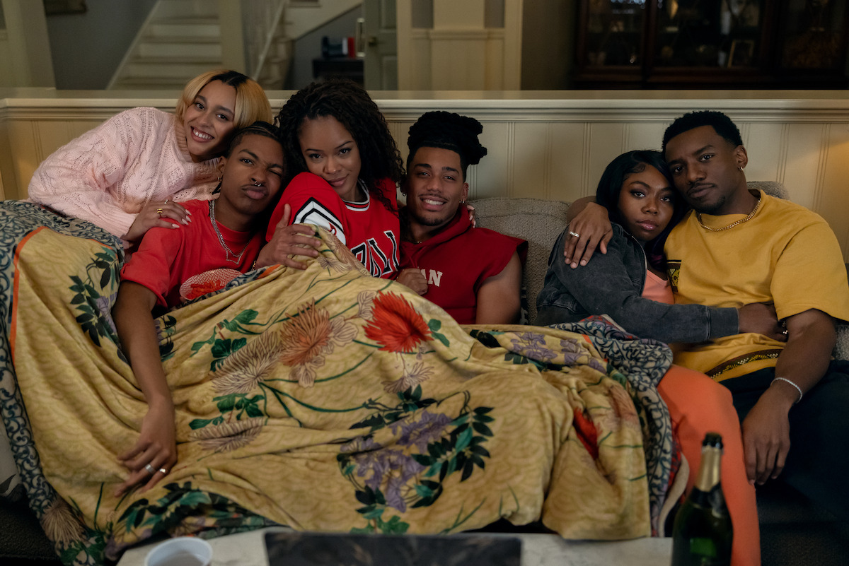 Six friends sitting closely together on a couch indoors, wrapped in a floral blanket, smiling and relaxed in a cozy living room with warm lighting and home decor in the background.