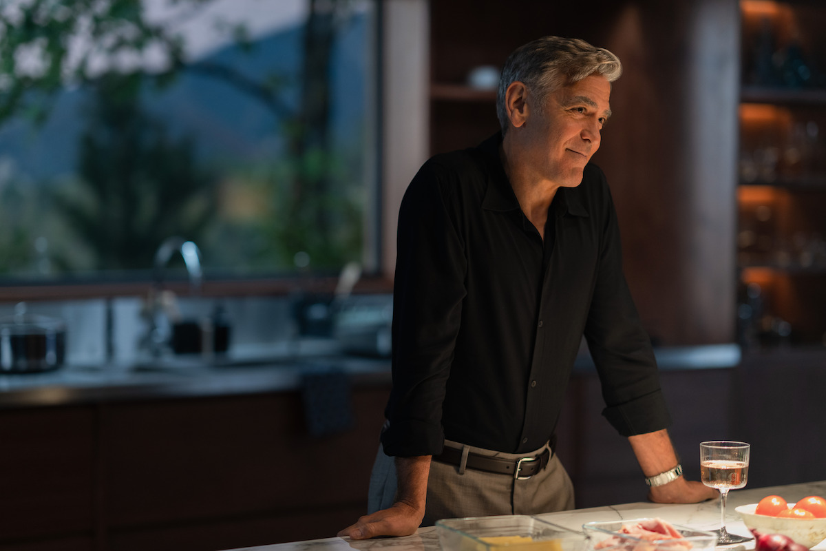 George Clooney with gray hair, wearing a black shirt, leans on a kitchen counter with a glass of wine and some food in front of him, while looking thoughtfully to the side.