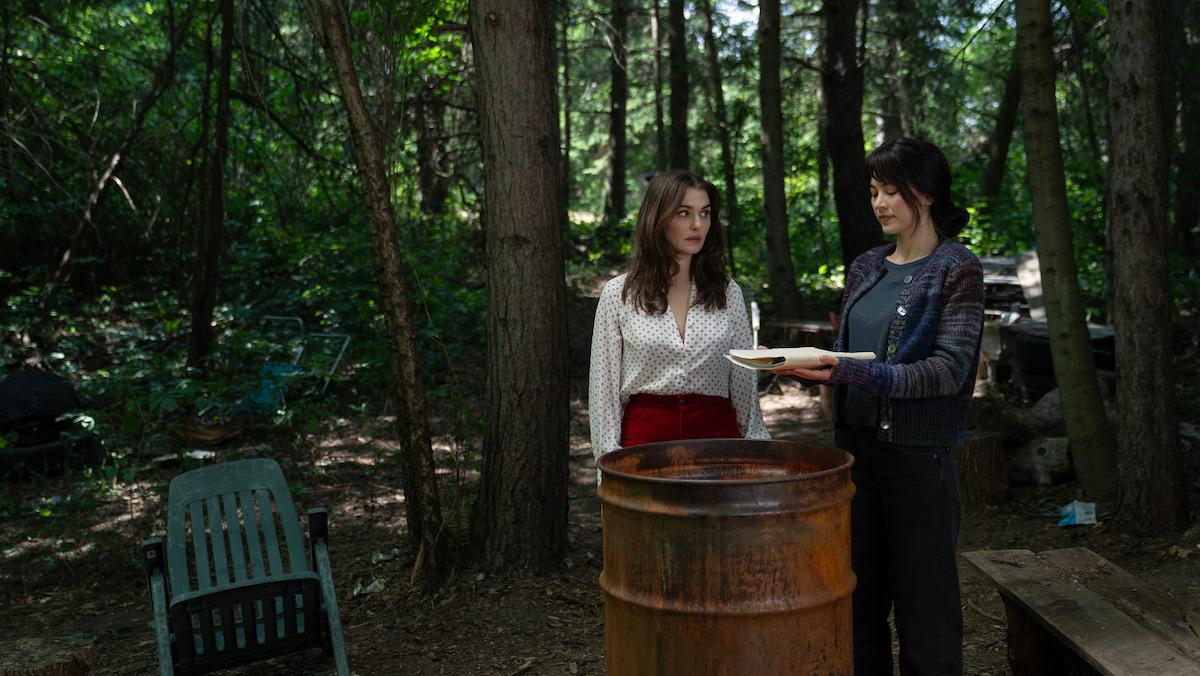 Rachel Weisz as The Protagonist and Jessica Henwick as Cynthia standing by a rusty barrel in a forest, surrounded by trees and outdoor furniture in a natural, wooded setting. One woman is holding a plate and they appear to be talking.
