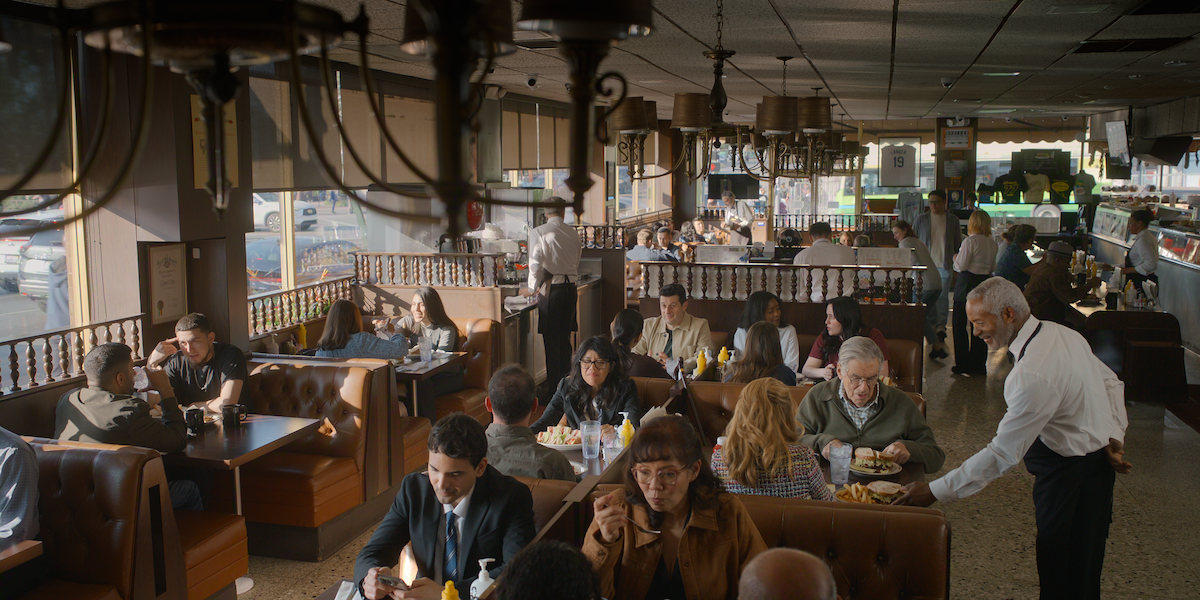 Busy diner interior with diverse diners eating at booths and tables, waiters serving food, ambient daylight coming through windows, and a classic retro atmosphere featuring brown seating and hanging light fixtures.
