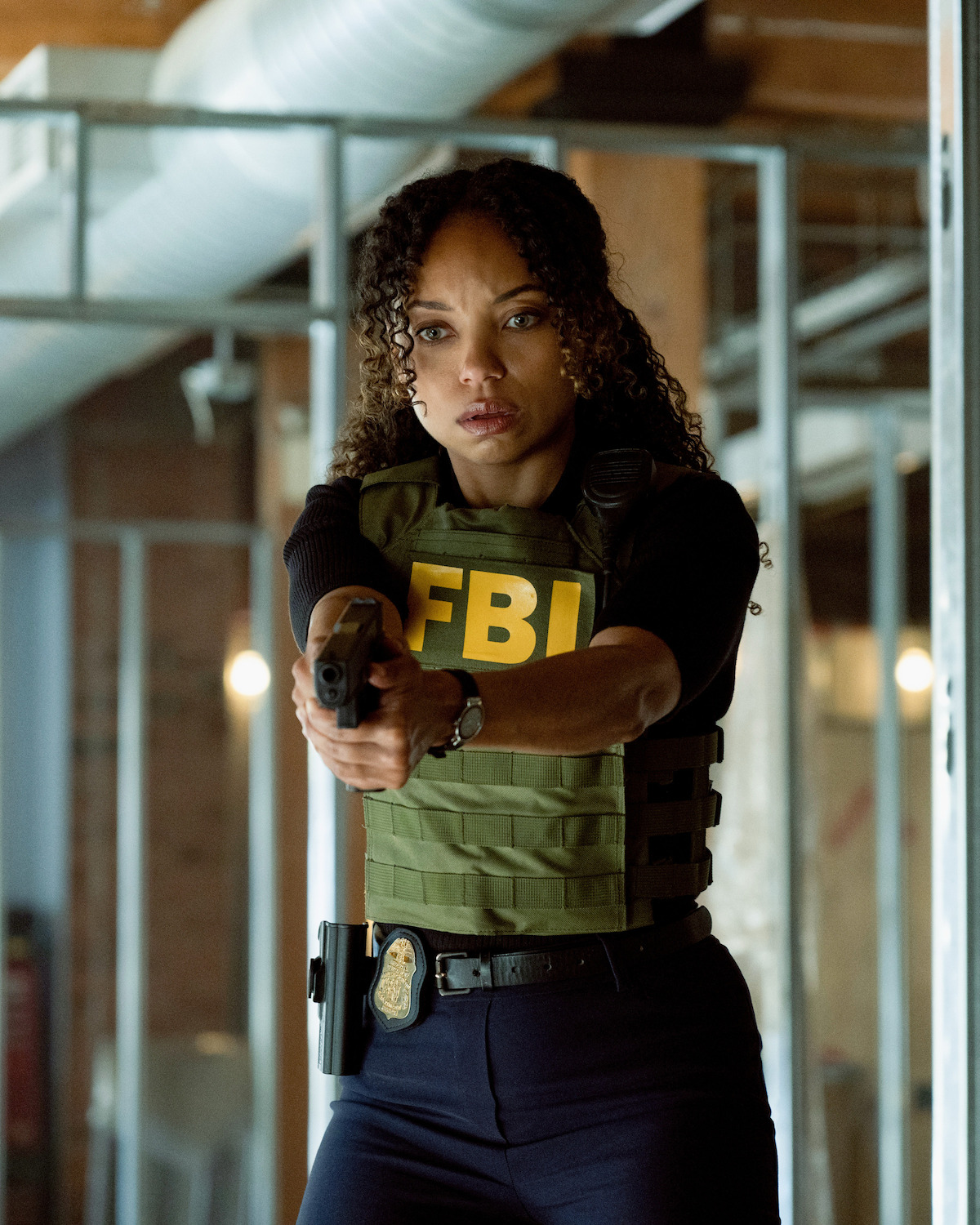 An FBI agent in a tactical vest aims her gun inside a building under construction with exposed metal framing, pipes, and brick walls, appearing alert and ready in a tense situation.