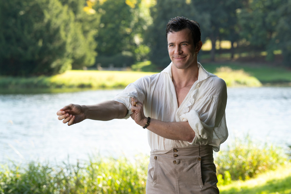 A man in period clothing sits confidently in a large leather armchair, adjusting his collar in a dimly lit, elegant room with candles on a nearby table, creating a sophisticated, historical atmosphere.