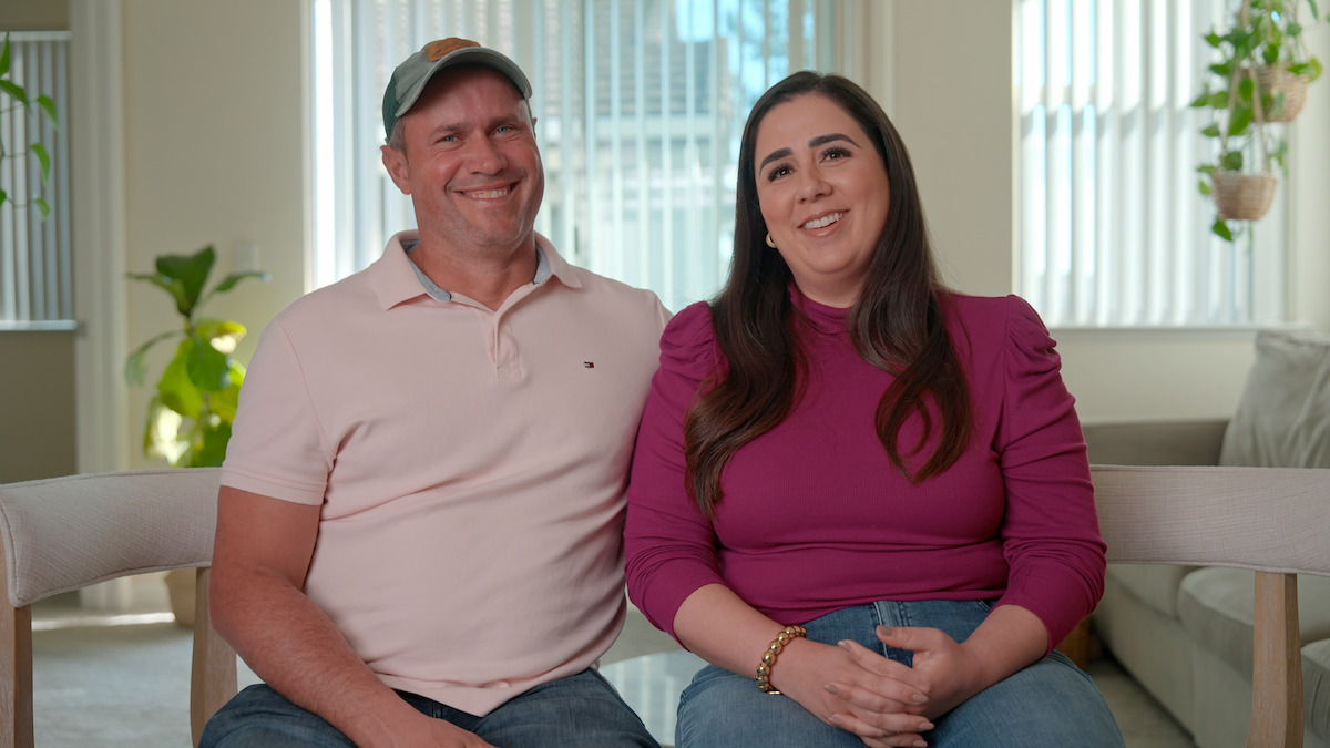 Smiling man in a light pink polo and cap sits next to a woman in a magenta top. They are indoors on a couch in a bright living room with plants and sunlight streaming through blinds in the background.