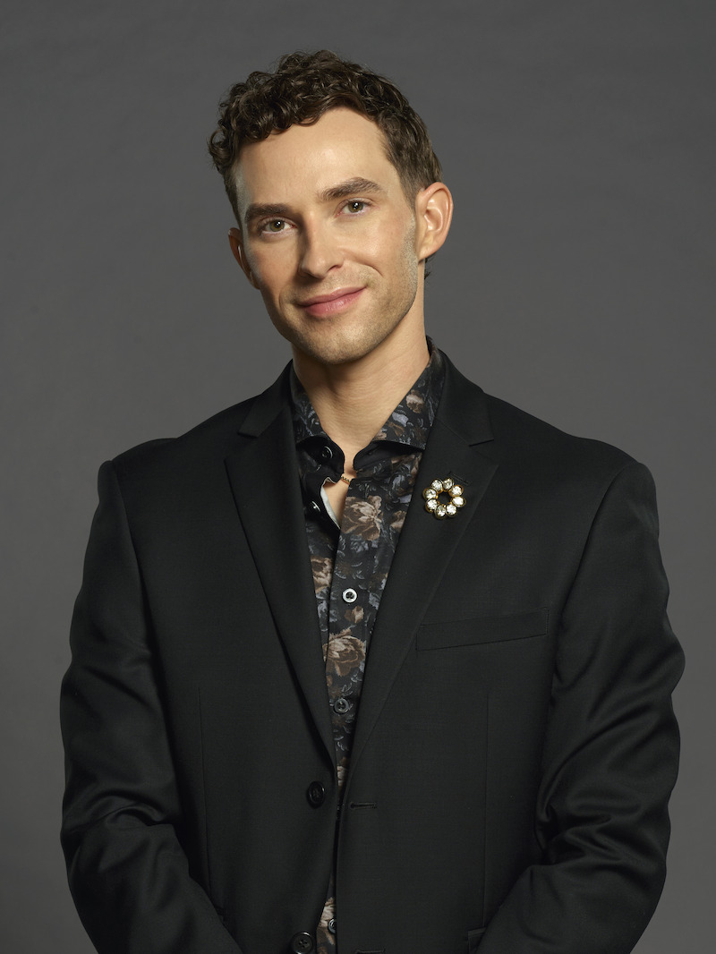 A person with short curly hair wearing a dark floral shirt and a black blazer with a brooch stands against a plain gray background, posing and smiling gently at the camera.