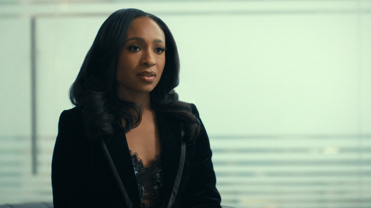 Woman with long dark hair in a black blazer sits indoors with a blurred glass wall and horizontal lines in the background, suggesting a modern office or professional setting.