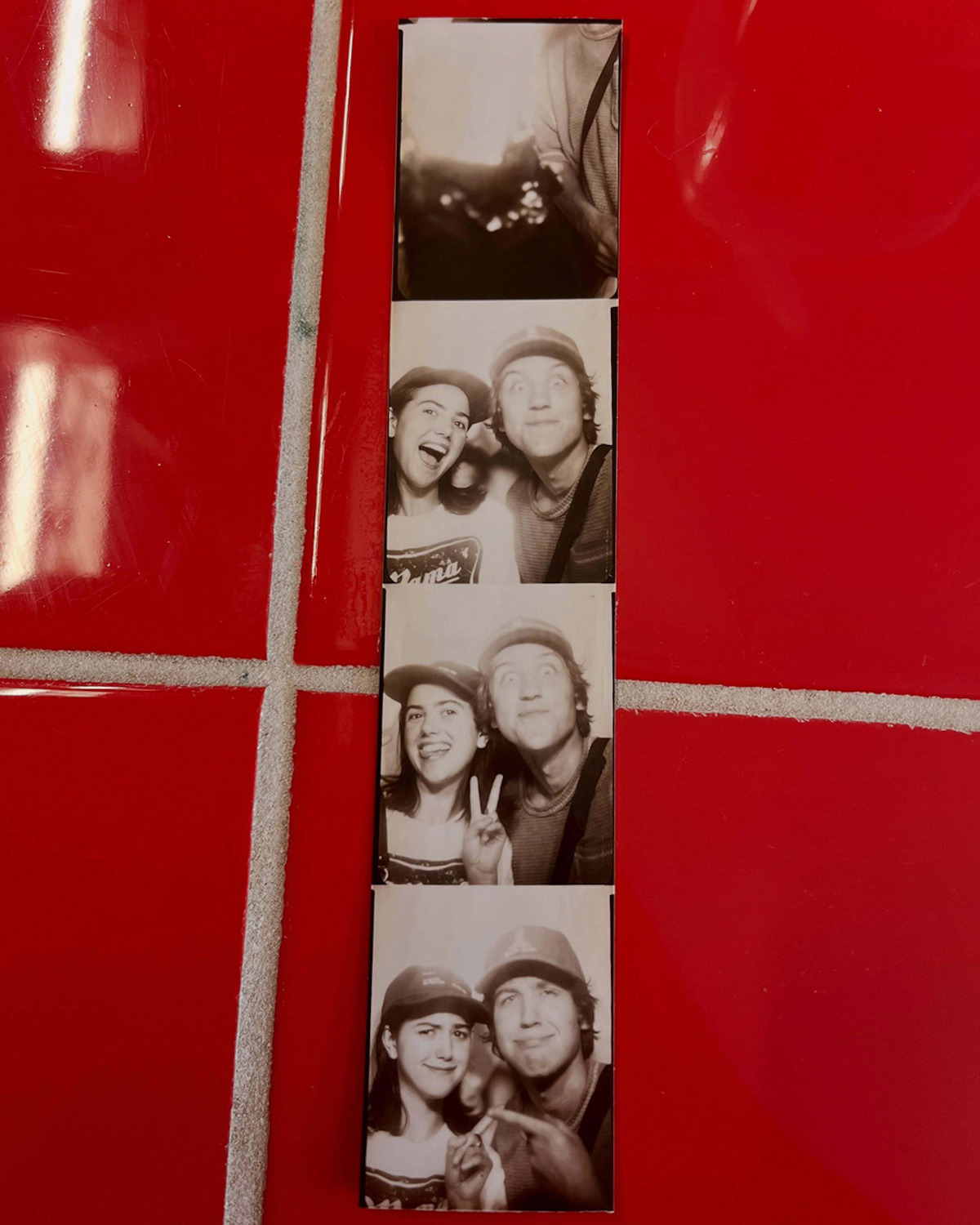 Black and white photo booth strip of two people making faces and peace signs, placed on a bright red tiled surface with white grout lines.